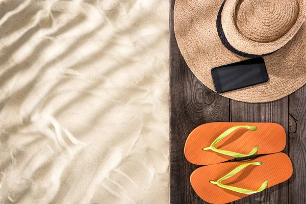 Vista superior de areia com espaço de cópia e chapéu de palha com fita preta, smartphone e chinelos laranja em placa marrom de madeira — Fotografia de Stock