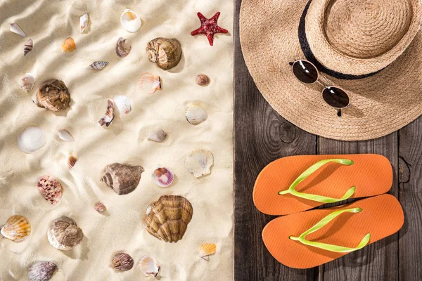 Top view of seashells and starfish on sand with orange flip flops, straw hat and sunglasses on wooden brown board — Stock Photo