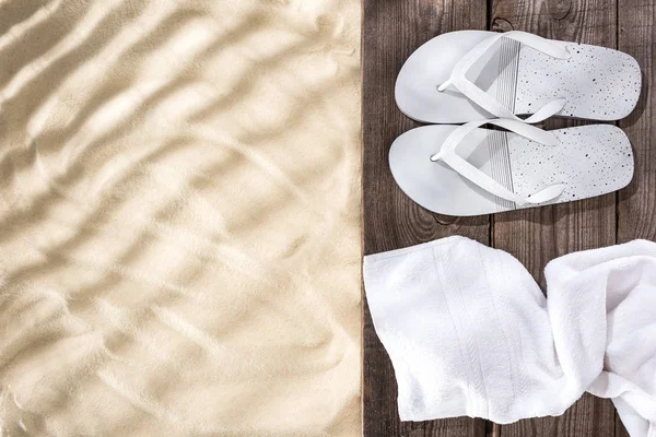 Vista superior de chinelos brancos e toalha na placa marrom de madeira e areia com espaço de cópia — Fotografia de Stock