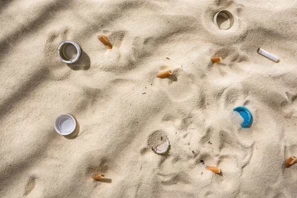 Vista dall'alto di conchiglie, tappi di bottiglia, mozziconi di sigaretta sparsi, tappi di plastica sulla sabbia — Foto stock