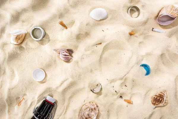 Vista dall'alto di conchiglie, tappi di bottiglia, mozziconi di sigaretta sparsi e bottiglia di vetro sulla sabbia — Foto stock