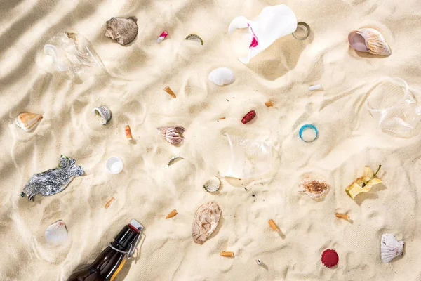 Top view of seashells, scattered cigarette butts, apple core, plastic cups, glass bottle and candy wrapper on sand — Stock Photo