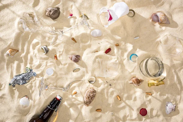 Vista dall'alto di conchiglie, bottiglia di vetro, mozziconi di sigaretta sparsi, vetri rotti, nucleo di mela, bicchieri di plastica e involucro di caramelle sulla sabbia — Foto stock