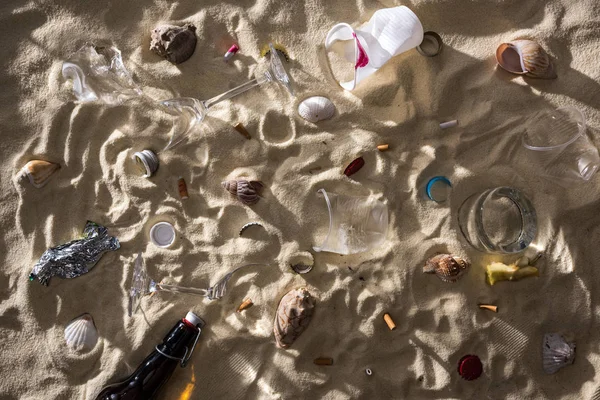 Vista dall'alto di conchiglie, bottiglia di vetro, mozziconi di sigaretta sparsi, vetri rotti, nucleo di mela, bicchieri di plastica e involucro di caramelle sulla sabbia con ombre — Foto stock