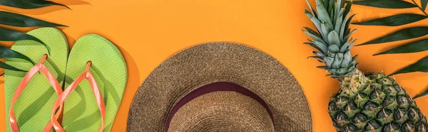 Panoramic shot of pineapple, tropical leaves, green flip flops and brown straw hat on orange background — Stock Photo