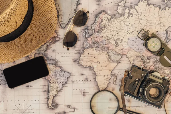 Top view of straw hat, sunglasses, film camera, magnifier, compass and smartphone with blank screen on world map — Stock Photo