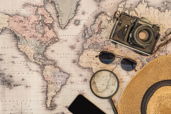 Vista superior de sombrero de paja, gafas de sol, cámara de cine, lupa y teléfono inteligente con pantalla en blanco en el mapa del mundo - foto de stock