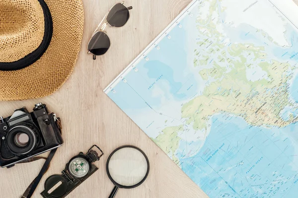 Top view of sunglasses, straw hat, compass, magnifier, film camera and world map on wooden surface — Stock Photo