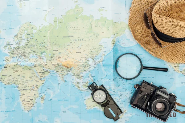 Vista superior del sombrero de paja, brújula, lupa y cámara de película en el mapa del mundo - foto de stock