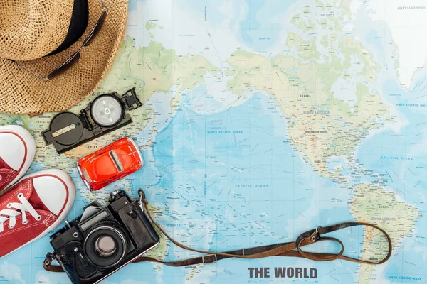 Vista superior de zapatos de goma, sombrero de paja, cámara de cine, gafas de sol. coche de juguete y brújula en el mapa del mundo - foto de stock