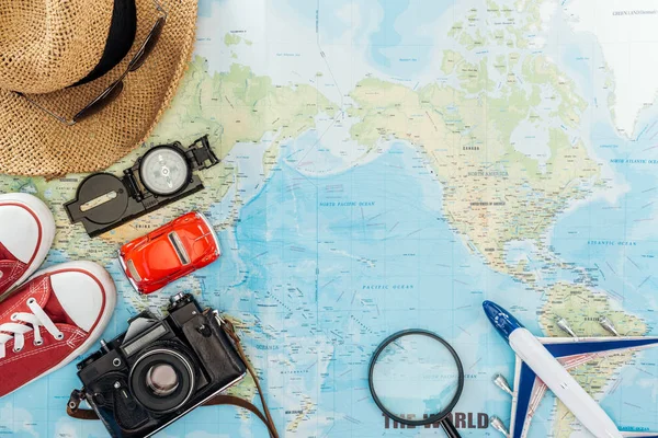 Visão superior da câmera de filme, chapéu de palha, óculos de sol, gomas, bússola, lupa, carro de brinquedo e avião de brinquedo no mapa do mundo — Fotografia de Stock