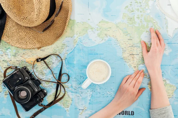 Vista recortada de la mujer con taza de capuchino, cámara de cine, gafas de sol y sombrero de paja en el mapa del mundo - foto de stock