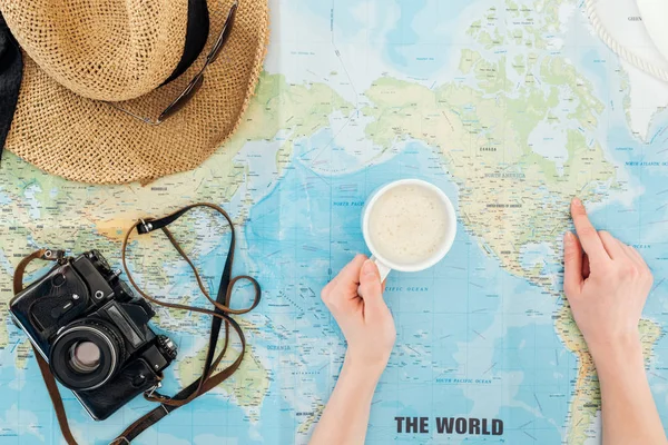 Vista recortada de la mujer con taza de capuchino, cámara de cine, gafas de sol y sombrero de paja que apunta con el dedo en el mapa del mundo - foto de stock