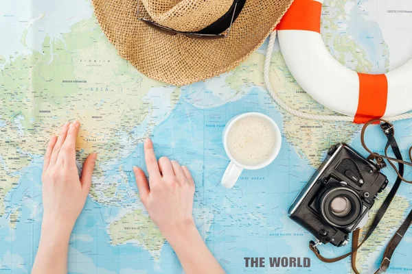 Vista recortada de mulher com xícara de cappuccino, câmera de filme, óculos de sol, boia salva-vidas e chapéu de palha apontando com o dedo no mapa do mundo — Fotografia de Stock