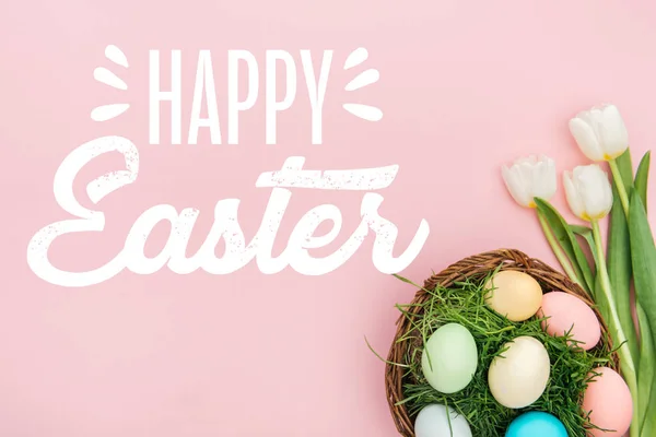 Top view of painted chicken eggs on green grass in wicker basket and white tulips on pink background with happy Easter lettering — Stock Photo