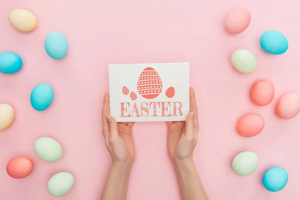 Cropped view of woman holding greeting card with Easter lettering near multicolored painted chicken eggs on pink background — Stock Photo