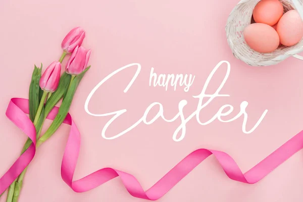 Vue du haut du bouquet de tulipes roses avec ruban et œufs peints dans un panier en osier avec joyeux lettrage de Pâques sur fond rose — Photo de stock