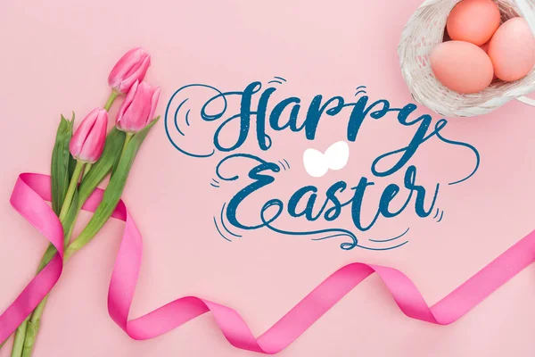 Top view of pink tulips bouquet with ribbon and painted eggs in wicker basket with happy Easter blue lettering on pink background — Stock Photo