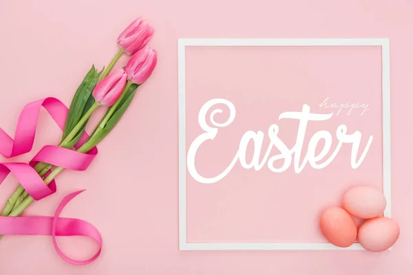 Ansicht von oben rosa Tulpenstrauß mit Schleife und bemalten Eiern in der Nähe Rahmen mit weißen glücklichen Ostern Schriftzug auf rosa Hintergrund — Stockfoto