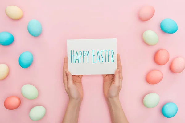 Partial view of woman holding greeting card with happy Easter lettering near multicolored painted chicken eggs on pink background — Stock Photo