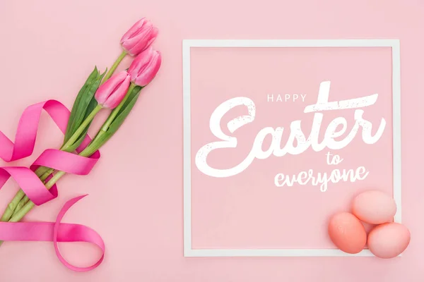 Top view of pink tulips bouquet with ribbon and painted eggs near frame with white happy Easter to everyone lettering on pink background — Stock Photo