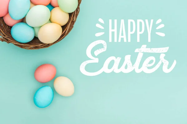 Top view of multicolored painted eggs in wicker basket with white happy Easter lettering on blue background — Stock Photo