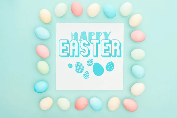 Top view of square frame made of painted chicken eggs on blue background with blue happy Easter lettering on white greeting card — Stock Photo