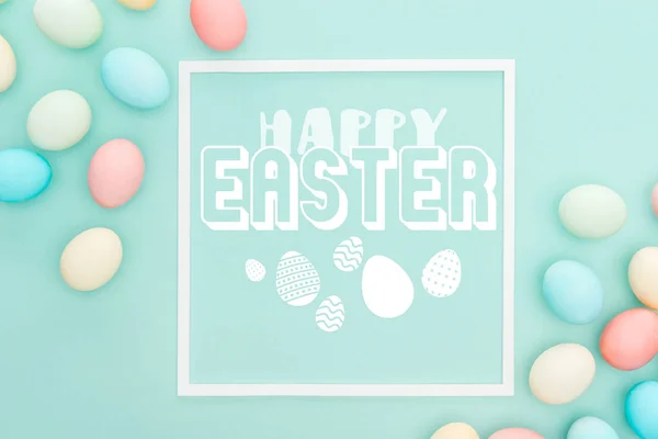 Top view of happy Easter lettering in white square frame near painted chicken eggs on blue background — Stock Photo