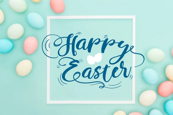 Top view of happy Easter blue lettering in white square frame near painted chicken eggs on blue background — Stock Photo