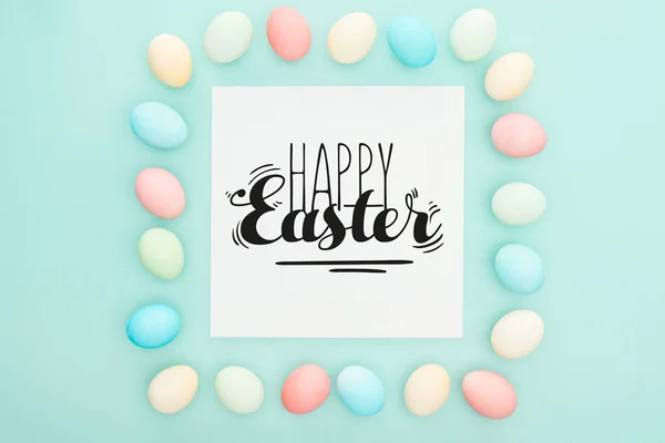 Top view of square frame made of painted chicken eggs on blue background with happy Easter black lettering on white greeting card — Stock Photo