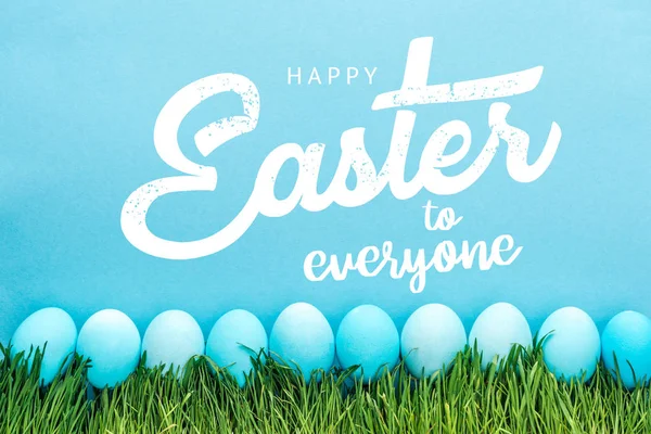 Blue painted chicken eggs on green grass with happy Easter to everyone lettering on blue background — Stock Photo