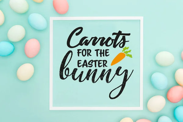 Top view of carrots for the Easter bunny lettering in white square frame near painted chicken eggs on blue background — Stock Photo