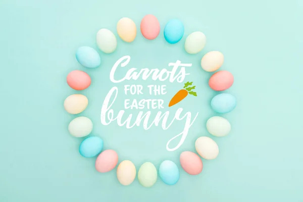 Top view of round frame made of painted chicken eggs on blue background with white carrots for the Easter bunny lettering — Stock Photo