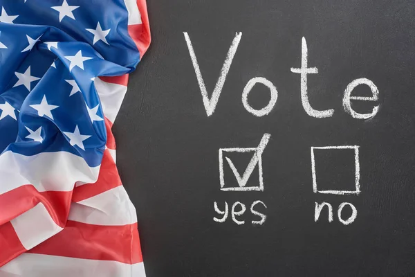 Top view of vote lettering and check mark near yes word on black chalkboard near american flag — стоковое фото