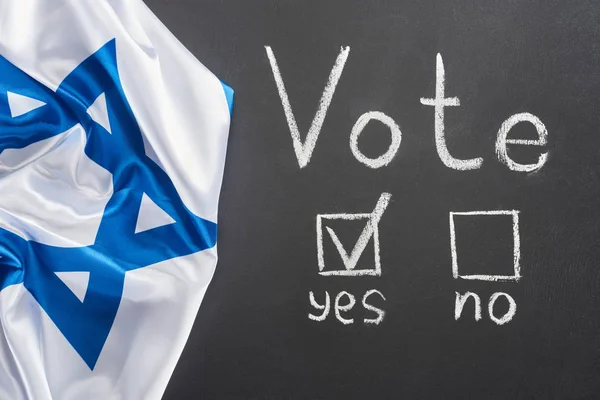 Top view of white vote lettering and check mark near yes word on black chalkboard near flag of Israel — стоковое фото