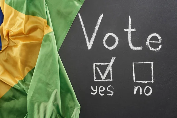 Top view of vote lettering and check mark near yes word on black chalkboard near flag of Brazil — стоковое фото