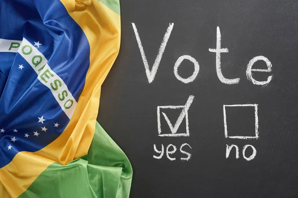 Top view of white vote lettering and check mark near yes word on black chalkboard near flag of Brazil — стоковое фото