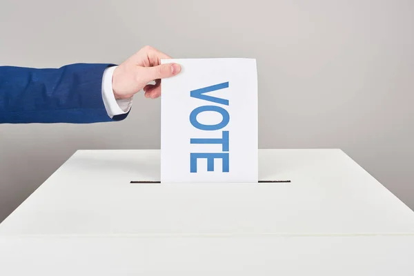 Cropped view of man putting card with vote in box on grey background — Stock Photo