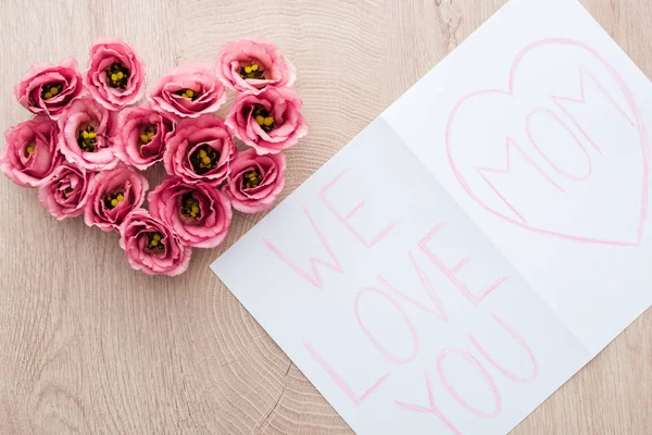 Vista superior de flores de eustoma en forma de corazón y tarjeta con nos encanta que mamá letras en la mesa — Stock Photo