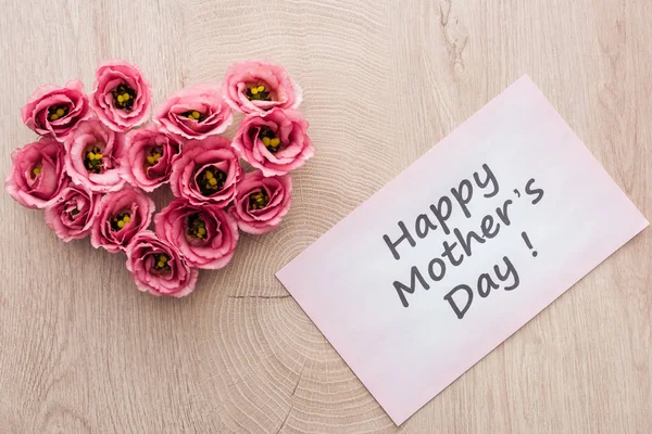 Vista dall'alto del segno del cuore fatto di fiori di eustoma e biglietto di auguri con felice giorno delle madri lettering sul tavolo di legno — Foto stock