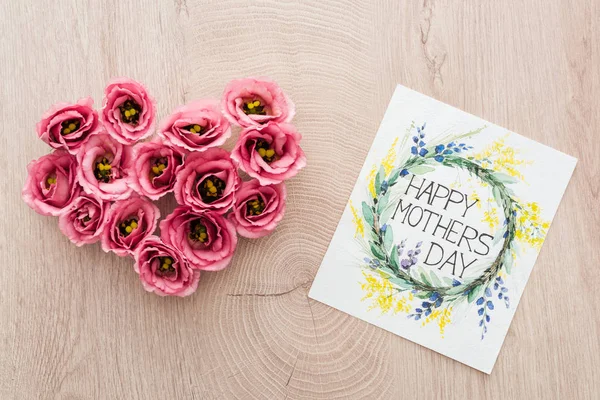 Vista superior del signo del corazón hecho de flores de eustoma y tarjeta con letras felices del día de las madres en la mesa de madera - foto de stock