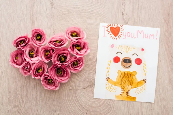 Top view of heart sign made of eustoma flowers and card with i love you mom lettering on wooden surface — Stock Photo