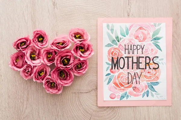 Top view of heart sign made of eustoma flowers and greeting card with happy mothers day lettering on wooden table — Stock Photo