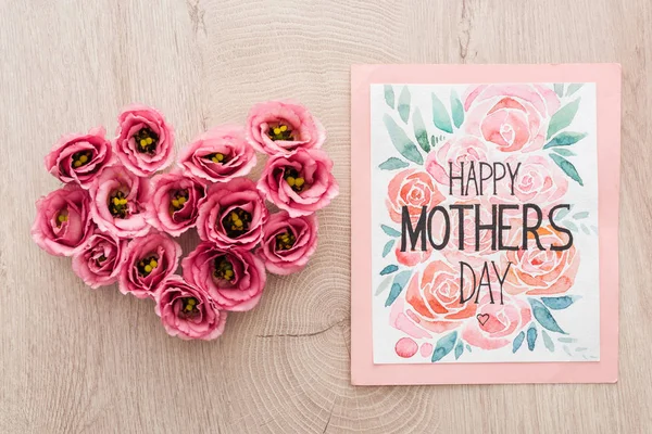 Vista dall'alto del segno del cuore fatto di fiori di eustoma e biglietto di auguri con felice giorno delle madri lettering sul tavolo di legno — Foto stock