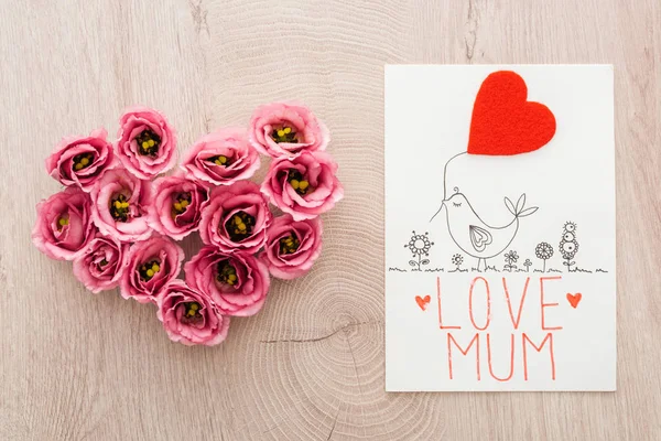 Vista dall'alto del segno del cuore fatto di fiori di eustoma e carta con amore mamma lettering su tavolo di legno — Foto stock