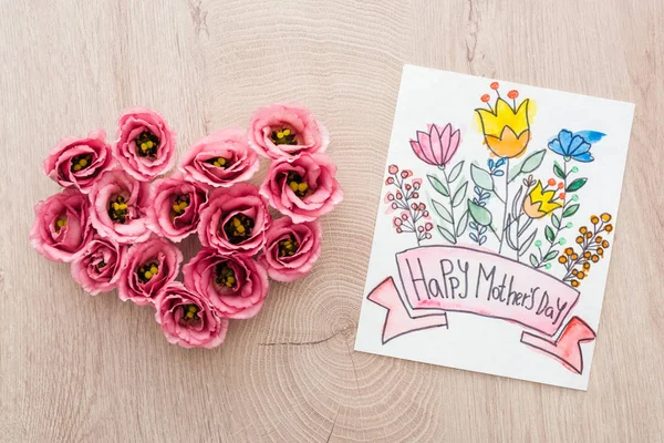 Vista superior del signo del corazón hecho de flores de eustoma y tarjeta con feliz día de las madres escribiendo en la mesa de madera - foto de stock