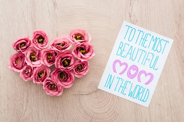 Top view of heart sign made of eustoma flowers and card with to the most beautiful mom in the world lettering on wooden table — Stock Photo