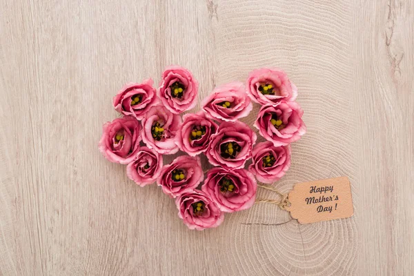 Vista superior del signo del corazón hecho de flores de eustoma con la etiqueta de papel del día de las madres felices en la mesa de madera - foto de stock