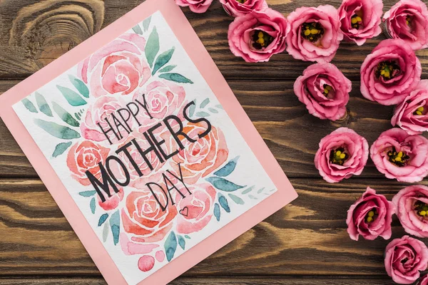 Vista superior de flores eustoma e cartão com feliz dia das mães lettering na mesa de madeira — Fotografia de Stock