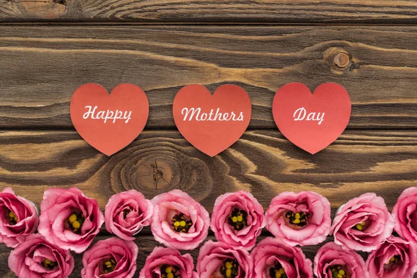 Vista dall'alto di fiori di eustoma rosa e cuori di carta rossa con scritte di giorno di madri felici su tavolo di legno — Foto stock
